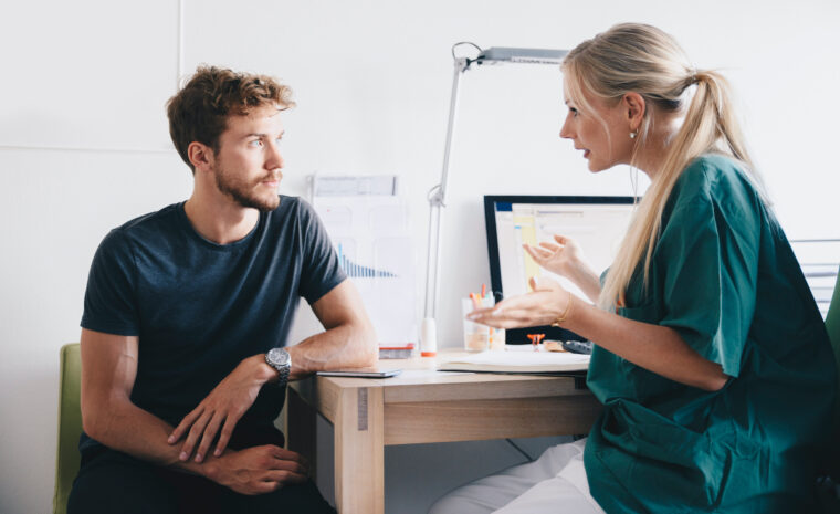 ung man lyssnar till kvinnlig vårdpersonal, de sitter vid kanten av ett skrivbord