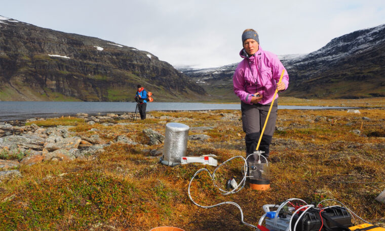 Kvinna i rosa jacka håller i mätinstrument. På myr med smält vatten. Fjäll och kollega i bakgrunden.