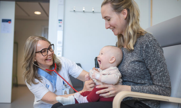 kvinnlig läkare skrattar mot spädbarn i mammas knä