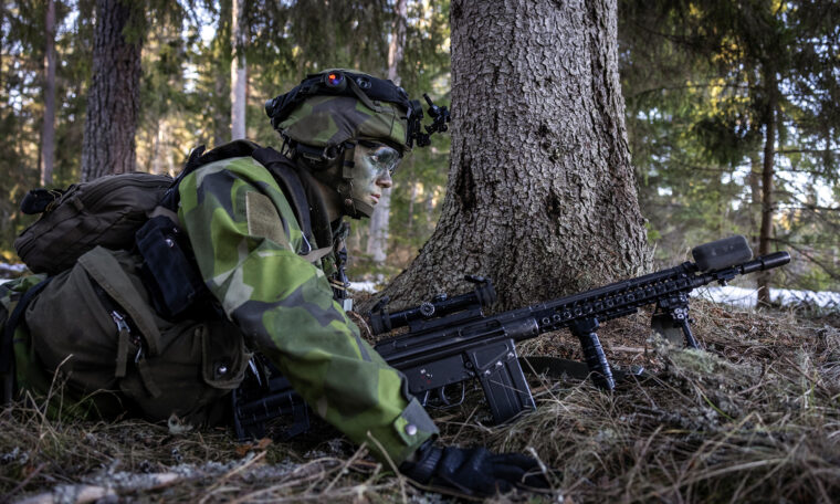 kamouflerad soldat ligger på marken bakom träd med vapen framför sig