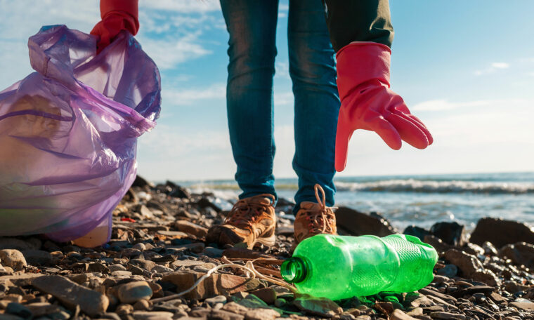 Person med röd skyddshanske plockar upp en grön plastflaska på stranden och lägger.